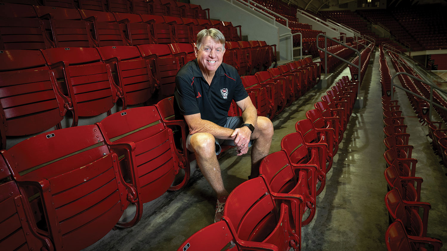 A portrait of a smiling Wes Moore, who sits in a row of empty seats at Reynolds Coliseum.