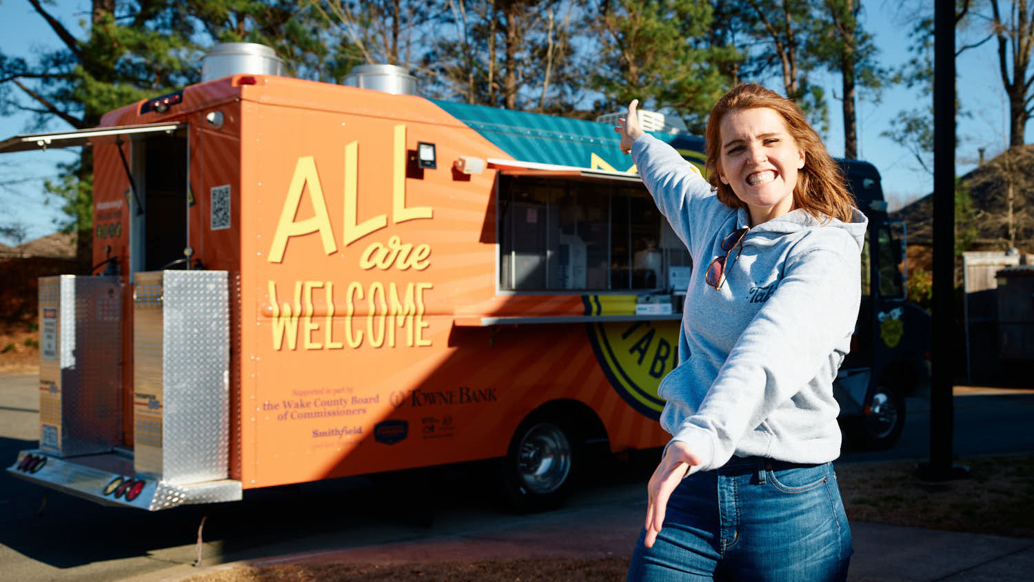 Maggie Kane stands in front of the Travelin' Table food truck. She smiles and gestures with her hands toward the truck, excited to show it off.