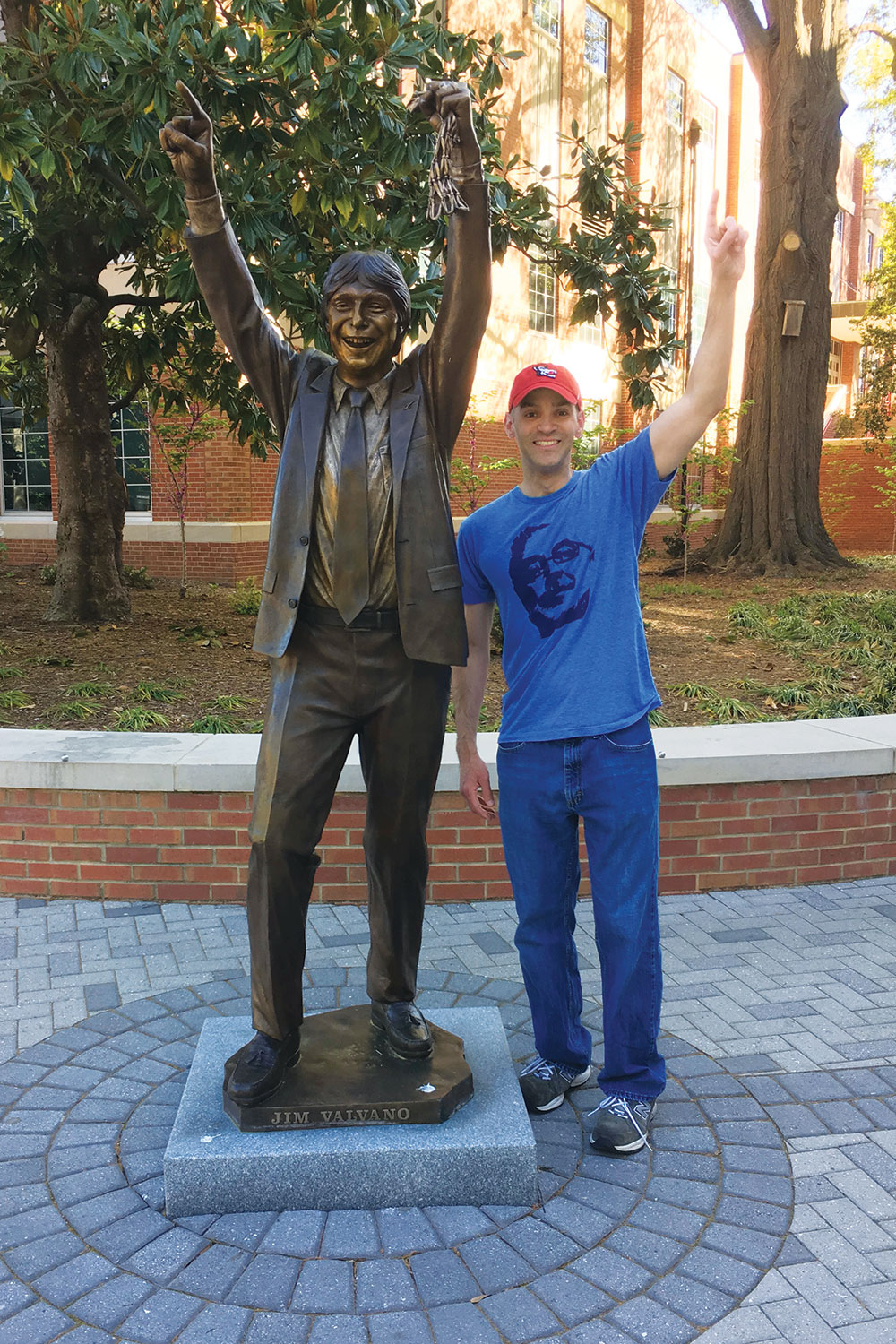 Lope “Max” Diaz II stands beside the Jim Valvano statue, smiling, mimicking Valvano's pose by making a number 1 sign with his hand and smiling.