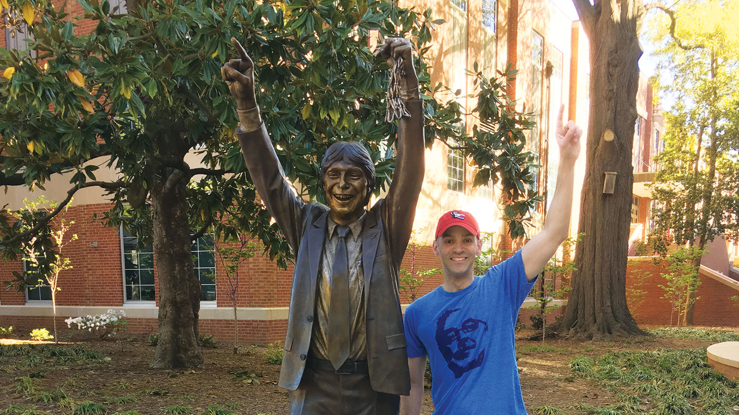 Lope “Max” Diaz II stands beside the Jim Valvano statue, smiling, mimicking Valvano's pose by making a number 1 sign with his hand and smiling.