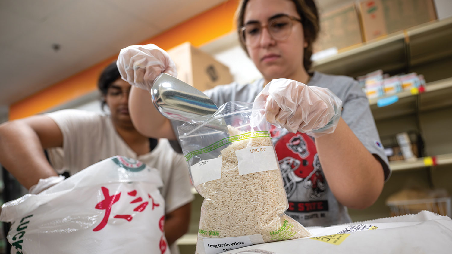 Two people wearing food-service gloves fill a zip-top bag of rice from a large bag or rice. In the background are boxes and shelves.