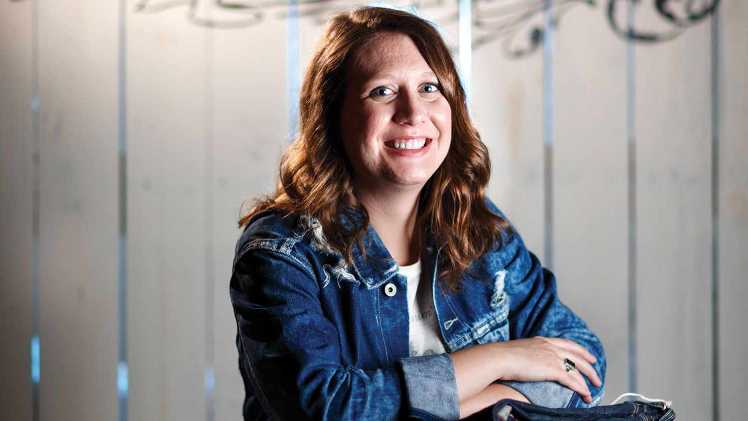 Caitlyn Holt, wearing a denim jacket and blue jeans, rests her folded arms on a stack of folded blue jeans.