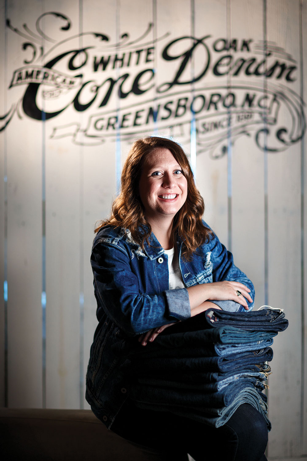 Caitlyn Holt, wearing a denim jacket and blue jeans, rests her folded arms on a stack of folded blue jeans.