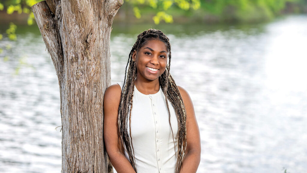 A portrait of Naomi Pitts leaning against a tree at the edge of Lake Raleigh.
