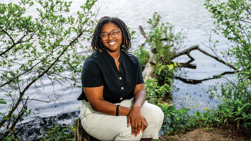 A portrait of Kayla Patterson sitting on a fallen tree at the edge of Lake Raleigh.
