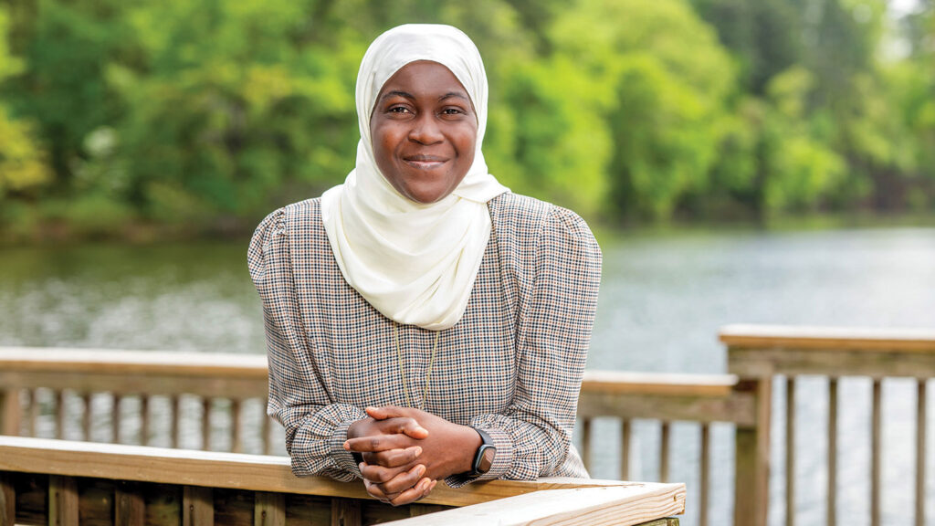 A portrait of Magreth Mushi, leaning on a wooden railing, in front of Lake Raleigh.