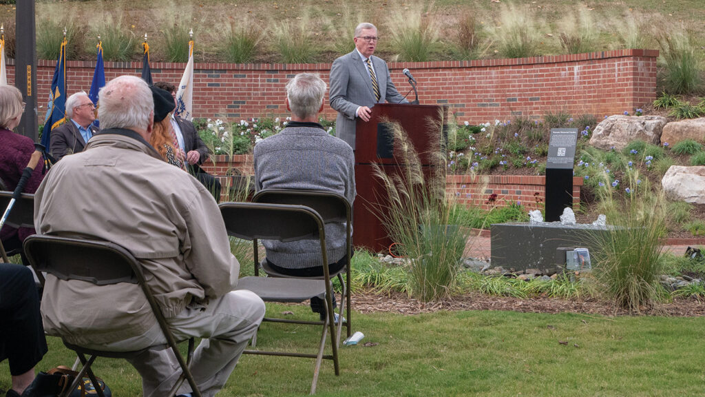 NC State Veterans Memorial Garden dedication ceremony with official speaking to audience near memorial fountain and reflection area surrounded by flowering plants and brick garden walls.