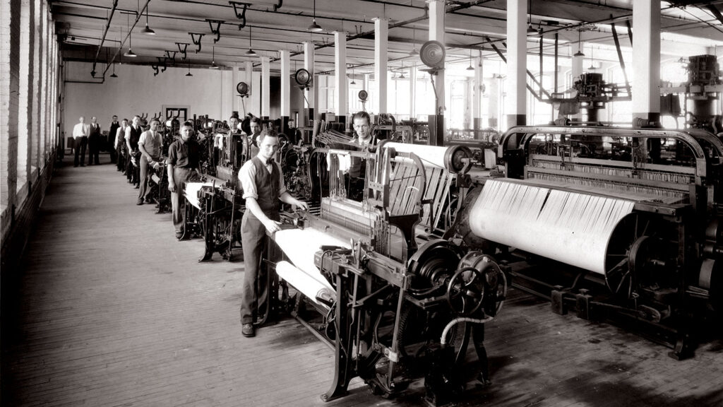 Students, dressed in button-down shirts, vests and ties, operate industrial textile looms in a 1930s College of Textiles facility. The black and white photo shows a large, bright room with rows of large weaving machines. It resembles an orderly factory floor.