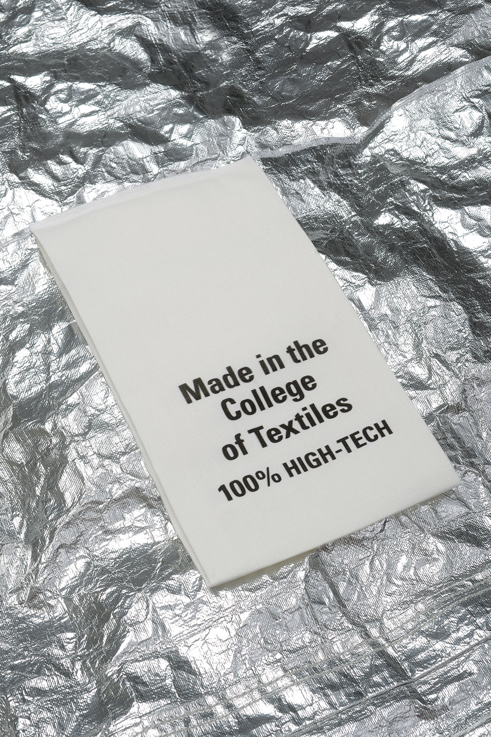 A white fabric care label reading "Made in the College of Textiles 100% HIGH-TECH" lies on a textured background of crumpled metallic silver material.