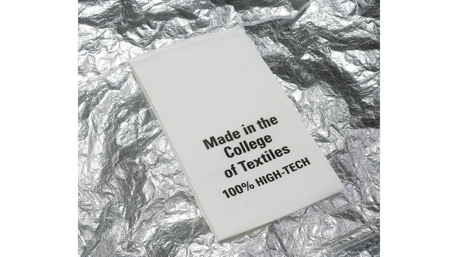 A white fabric care label reading "Made in the College of Textiles 100% HIGH-TECH" lies on a textured background of crumpled metallic silver material.