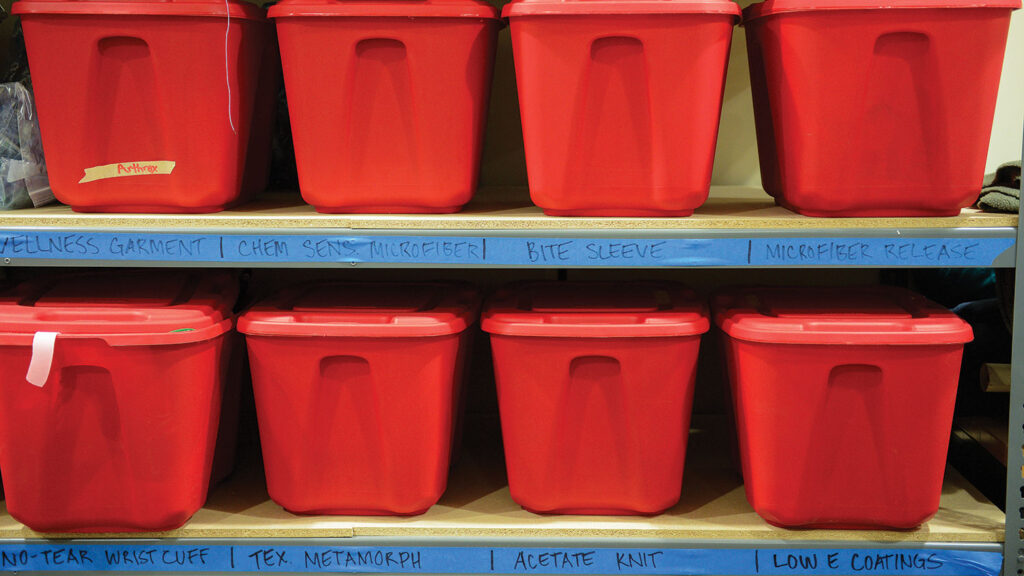 Red storage bins containing student textile projects in progress are organized on shelves with labels including "Wellness Garment," "Chem Sens Microfiber," "Bite Sleeve," "Microfiber Release," "No-Tear Wrist Cuff," "Tex Metamorph," "Acetate Knit" and "Low E Coatings."