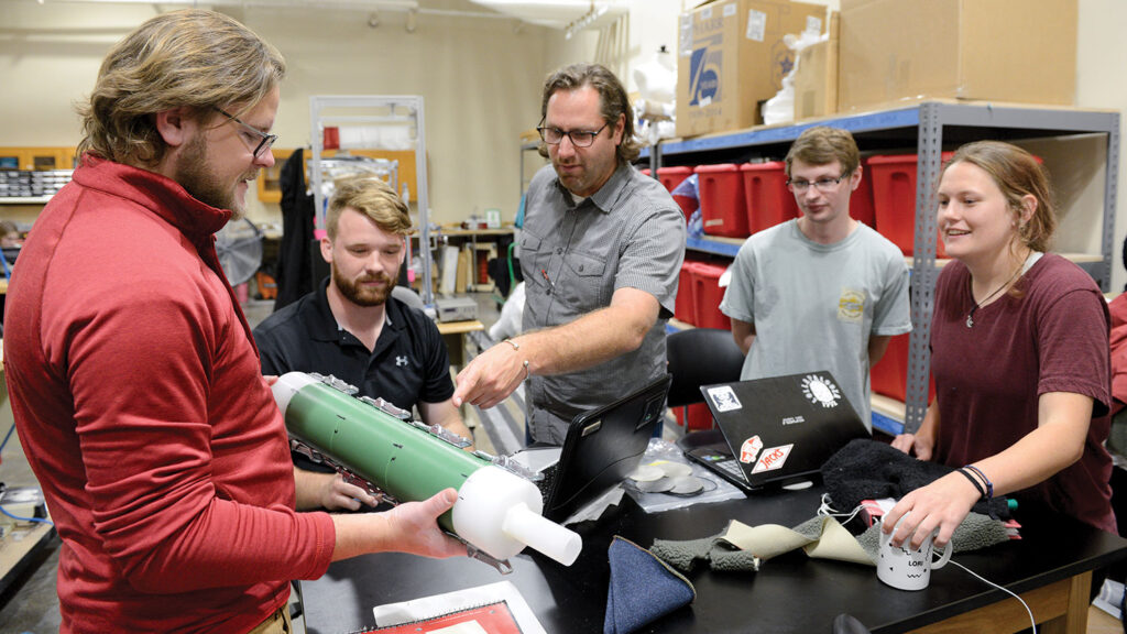 Two professors and three students gather around a table covered in fabric samples and laptop computers in a textiles laboratory. One of the professors points to a green cylindrical devise that the other is holding.