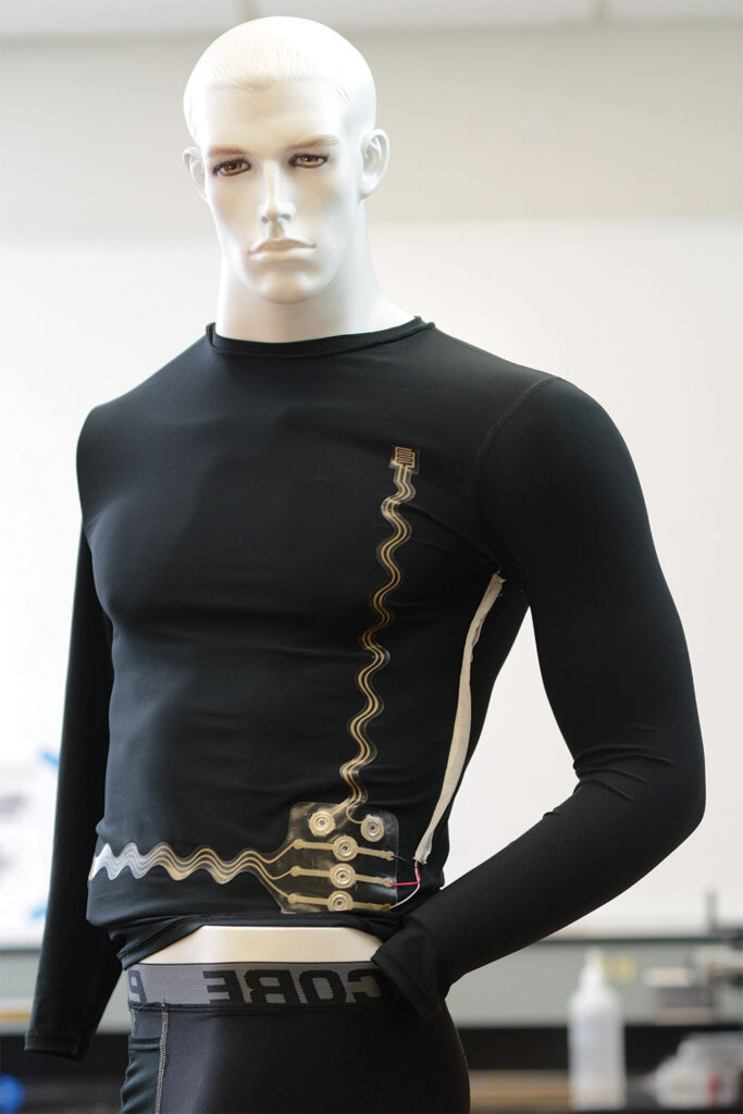 A mannequin displays a black long-sleeved shirt with gold conductive wires and electronic components integrated into the fabric, demonstrating high-tech, wearable textile technology.