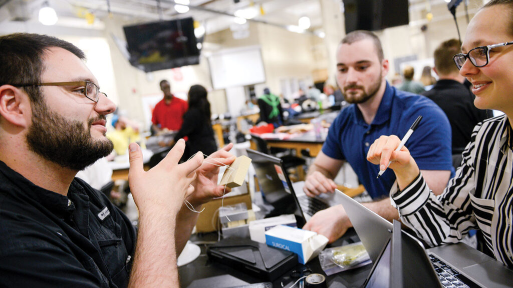 Three students collaborate in a textile laboratory. One person holds a small prototype object containing fiber strands. The other two students listen and take notes at a table with laptops and equipment.