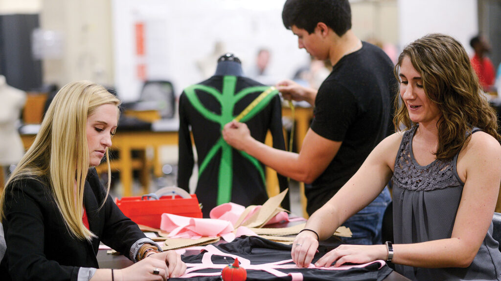 Two women work with fabric and patterns at a table in a textiles studio, while a man adjusts a green and black garment on a dress form in the background.