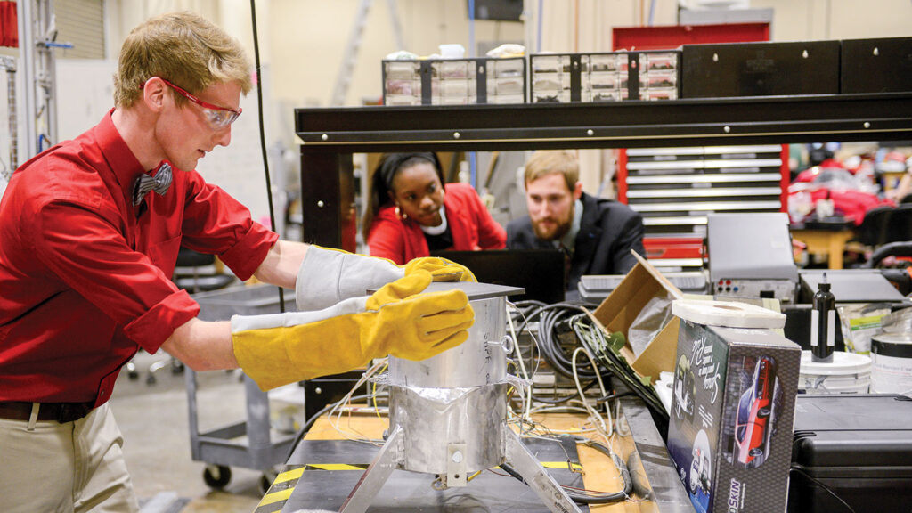 A student wearing safety glasses and yellow gloves operates laboratory equipment while two colleagues observe in a textile studio with other testing equipment visible in the background.