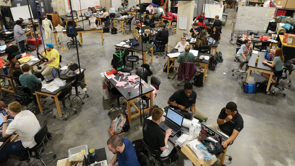 An overhead view of a busy textile engineering classroom showing approximately 20 students working at lab tables with laptops, equipment and materials. The industrial space has concrete floors and features a dress form mannequin, whiteboards and various textile testing equipment throughout.