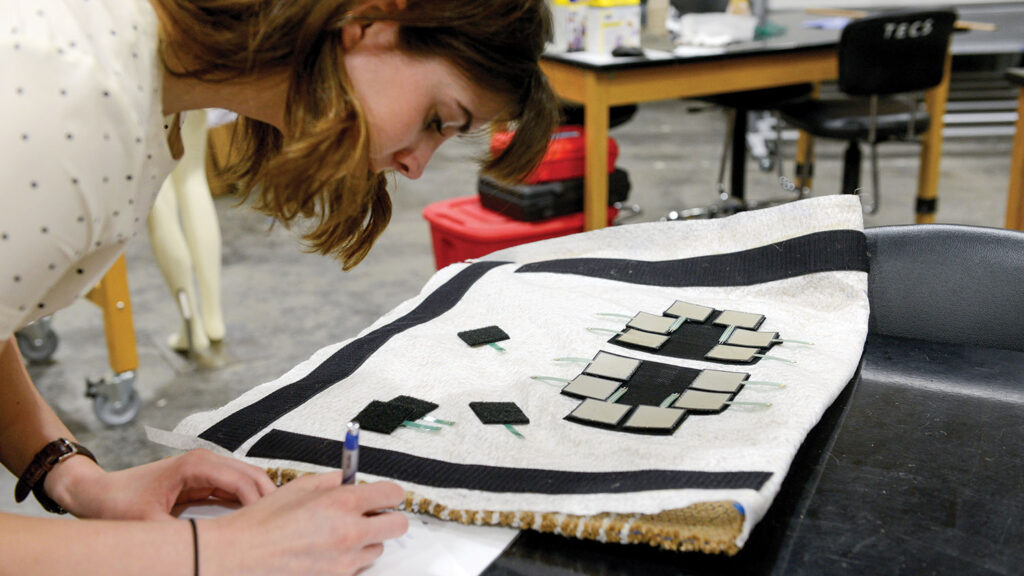 A student works on a bite sleeve. She has arranged black and white fabric patches in a geometric pattern on white material spread across a work table in a textile laboratory.