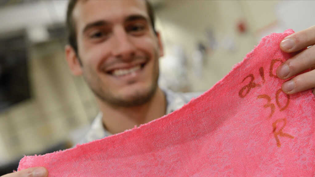 A smiling male student holds up a pink fabric swatch in a laboratory setting, with blurred textile equipment visible in the background.