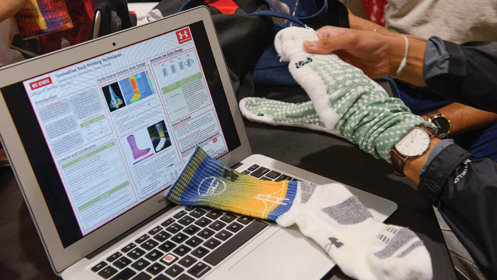 A student examines colorful patterned socks while a laptop displays research on "innovative sock printing techniques," with various textile samples and socks spread across the workspace.
