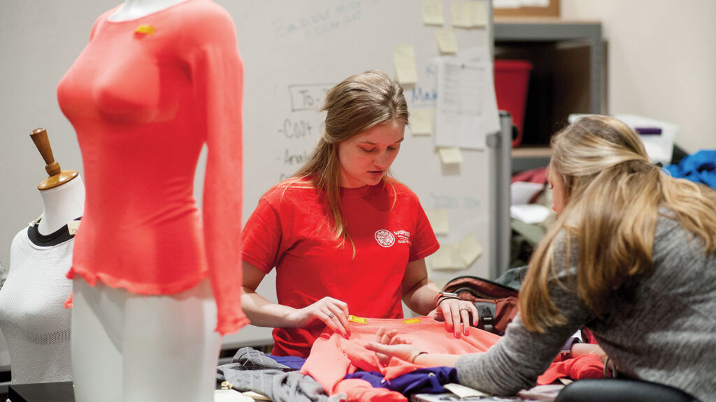 Two students work together to create an innovative T-shirt, with a bright coral-colored knitted garment displayed on a dress form in the foreground and fabric materials spread across their workspace.