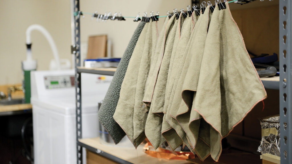 Samples of fleece fabric hang from clips on a metal rack after being run through an industrial washing machine to test microfiber release during laundering, with laboratory equipment visible in the background.