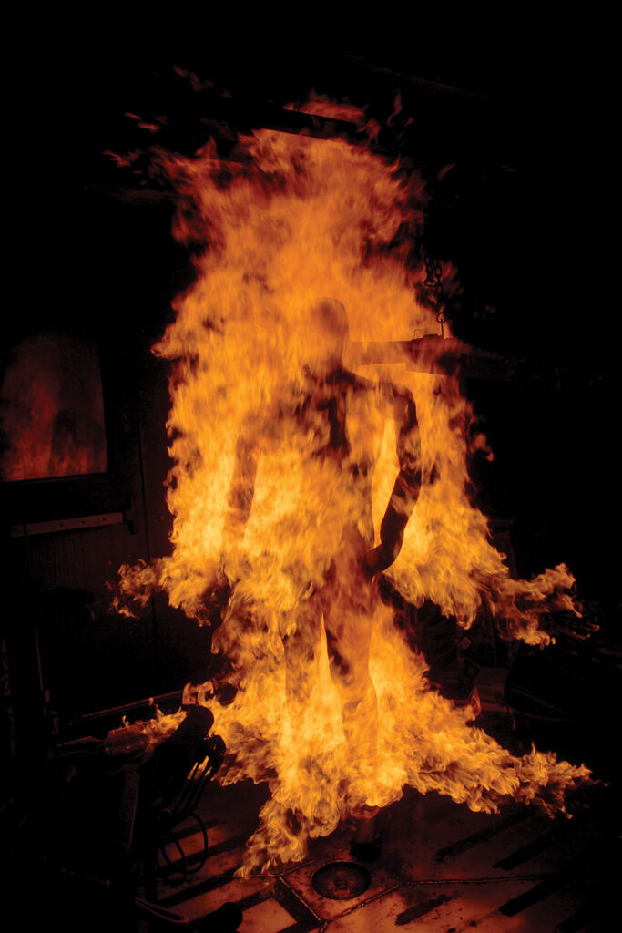 A mannequin called "Pyroman" wearing a fire-protective suit is engulfed in flames during a demonstration, with bright orange and yellow fire covering the entire figure against a dark background.