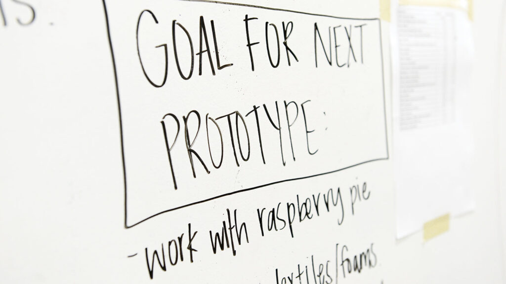 A handwritten sign on a whiteboard reads "GOAL FOR NEXT PROTOTYPE" with additional notes below including "work with raspberry pie" and partial text about "textiles/foams."